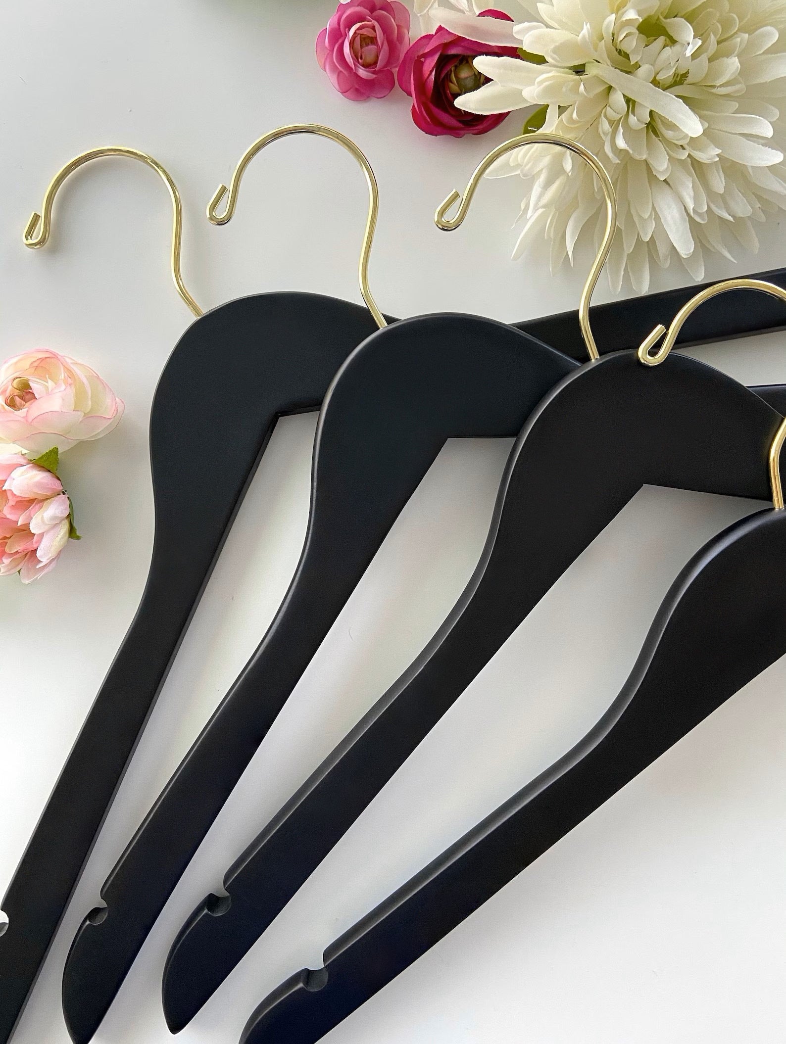 Four Matte Black Wooden Clothes Hangers with gold hooks for closets and stores lying on a table with flowers on it