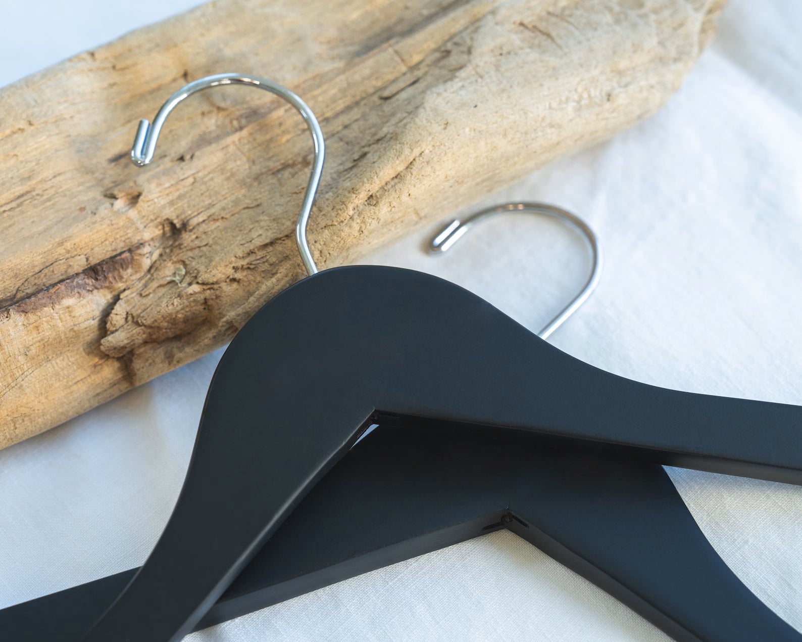 Two customizable Matte Black Wood Suit Hangers with silver hooks for adults lying down against a piece of wood on a table